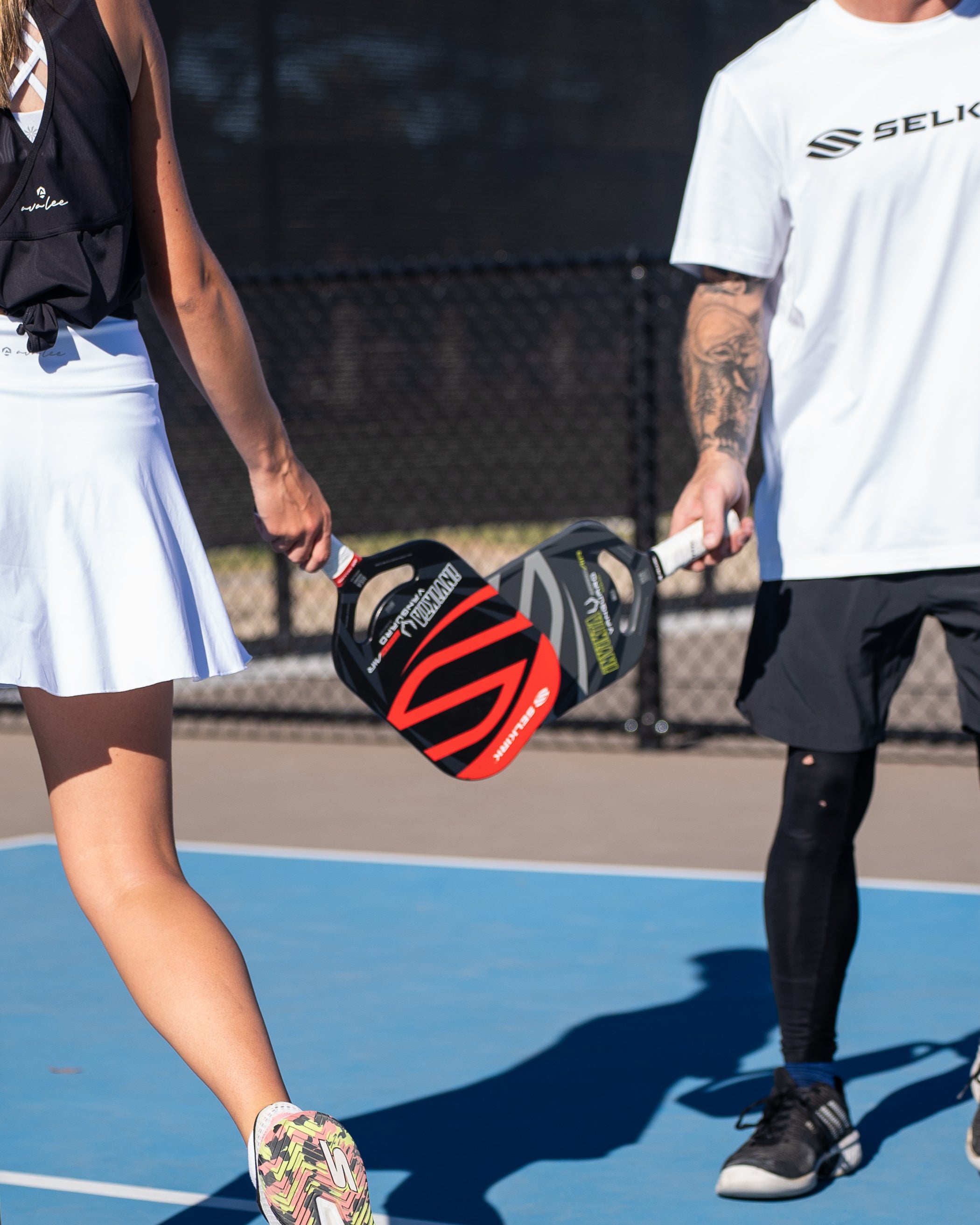 A man and a woman tap their coordinating Selkirk VANGUARD Power Air pickleball paddles on an outdoor pickleball court. 