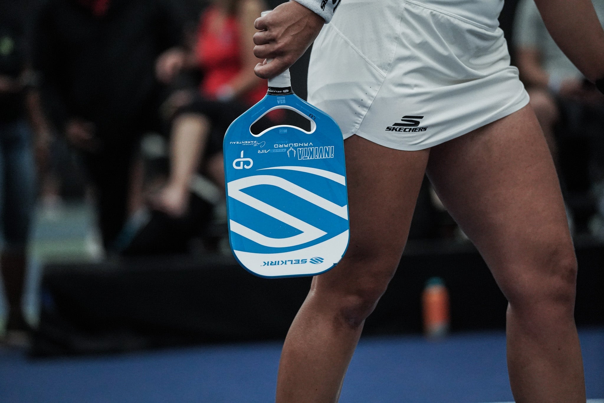 A woman holds a blue Selkirk VANGUARD Power Air, with an air dynamic throat, as she walks off court. 