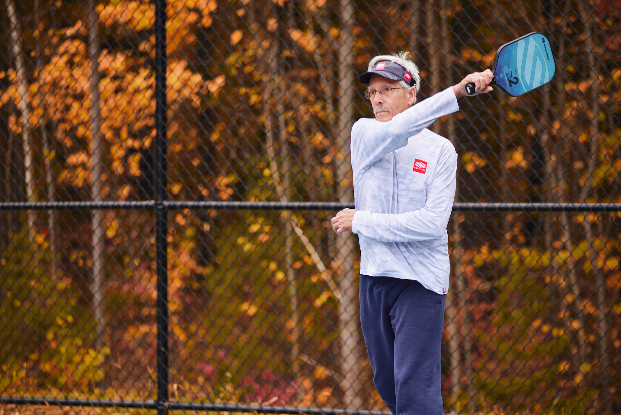 An older man wears long-sleeved layers as he plays pickleball outside. Behind him, the leaves are turning orange. 