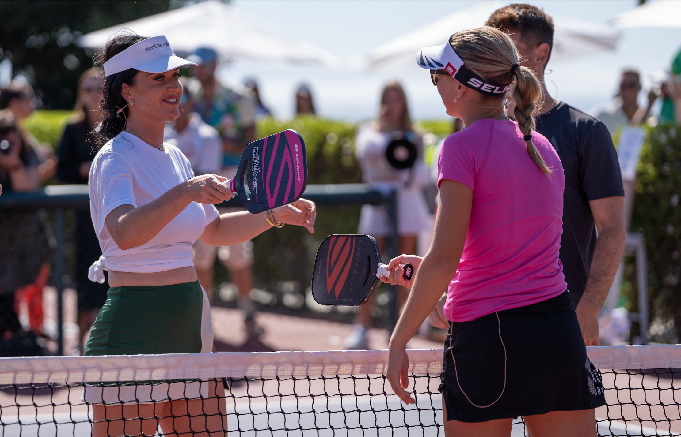 Katy Perry taps her Selkirk Power Air paddle with Selkirk pro Maggie Brascia over the net of an outdoor pickleball court. 