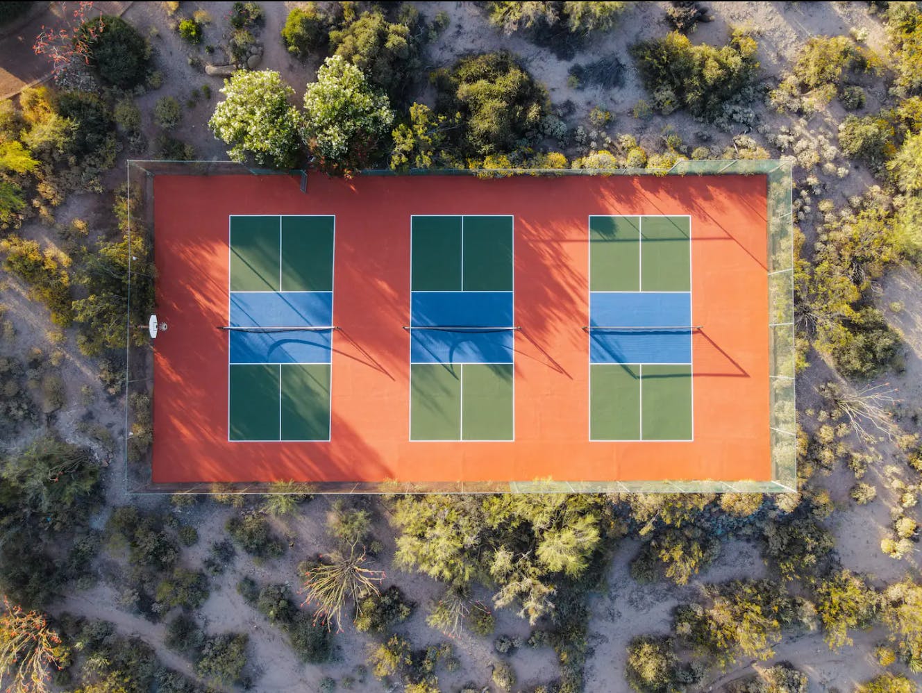 An aerial image shows three pickleball courts from above. They are situated in a desert. 