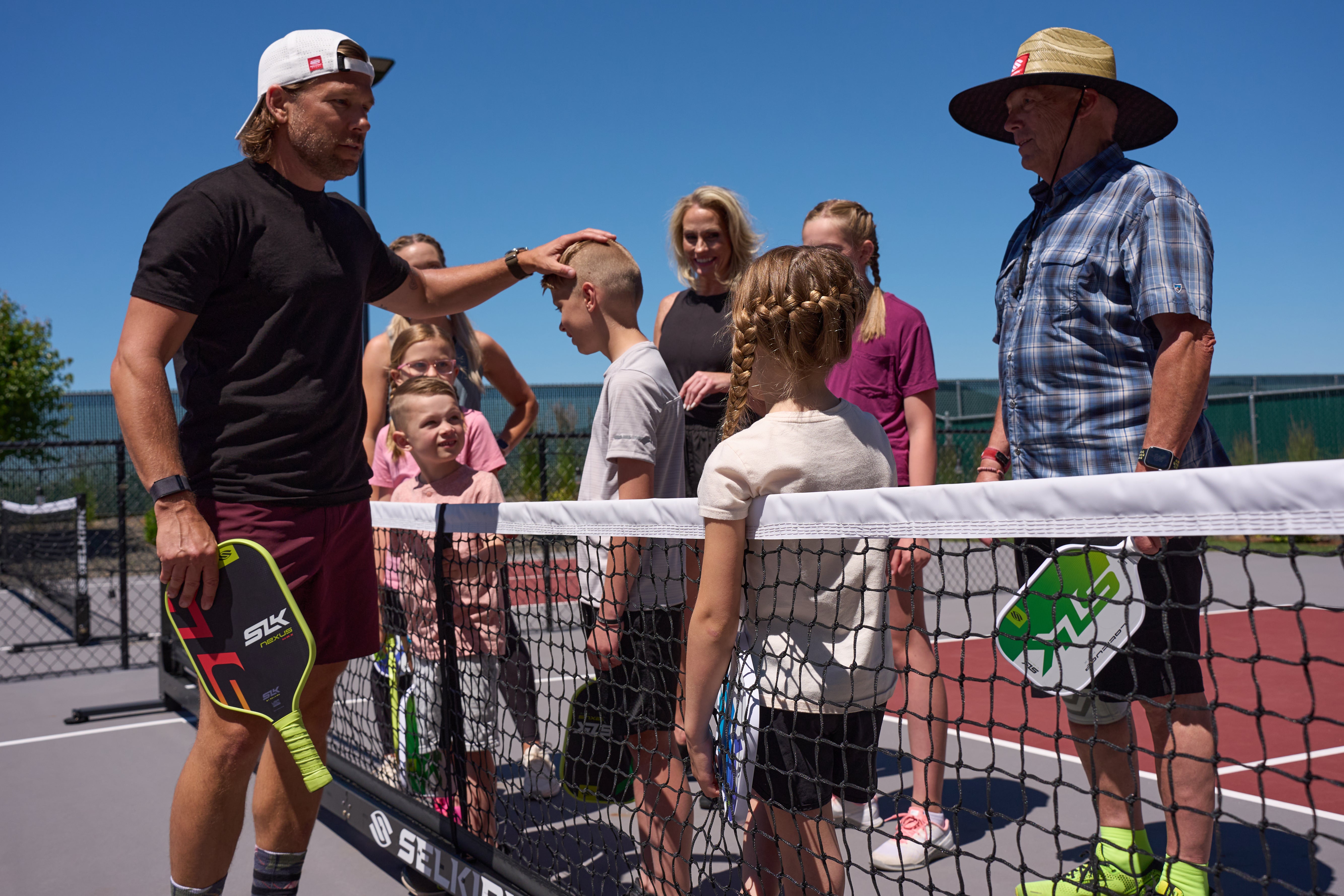 Children and adults of all ages smile as they gather near a the net on an outdoor pickleball court. 