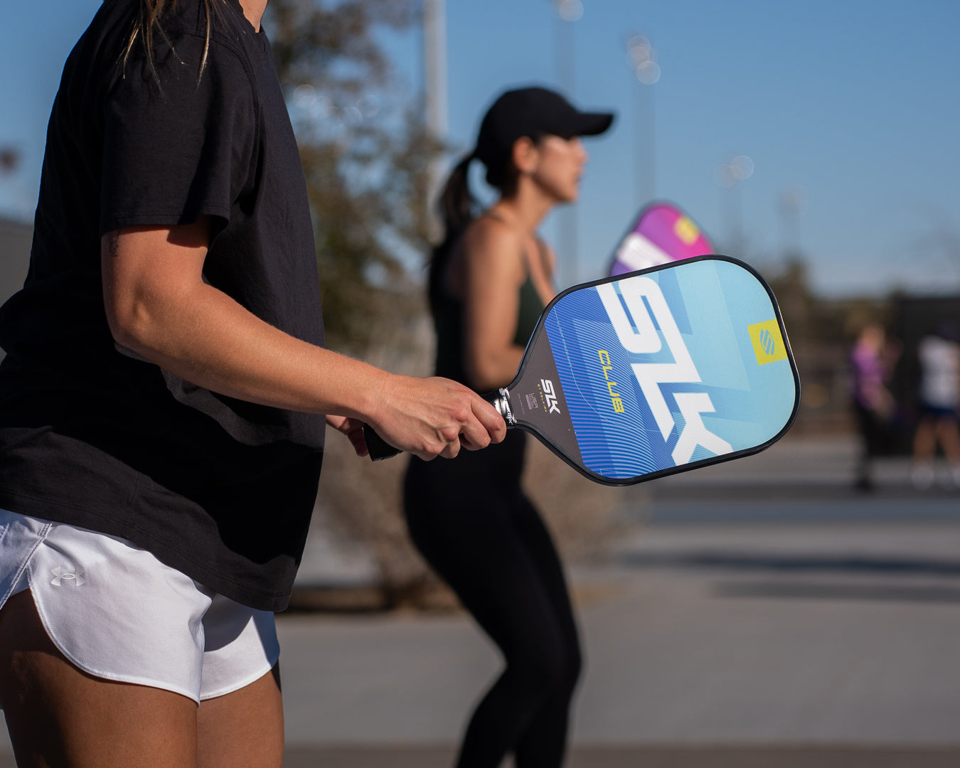 Two women prepare to receive a shot at the baseline of an outdoor pickleball court. 