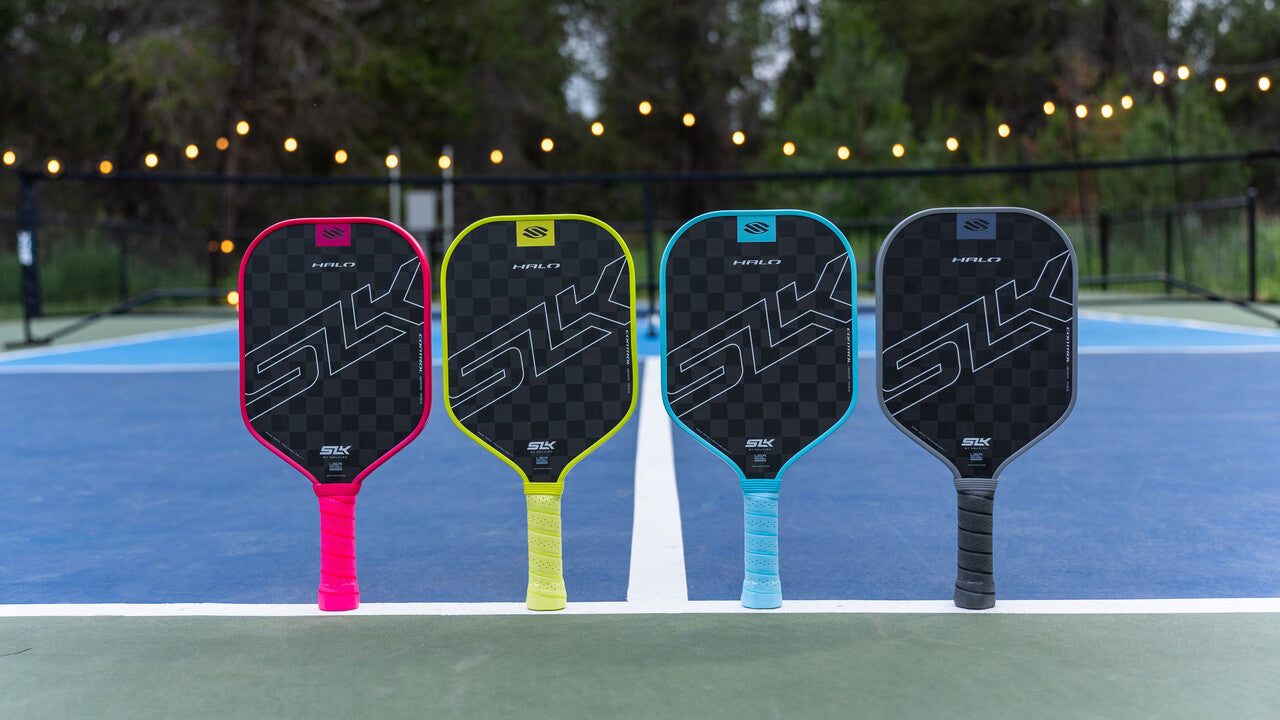 Four SLK Halo paddles stand vertically on their handles. They are lined up on the sideline of an outdoor pickleball court