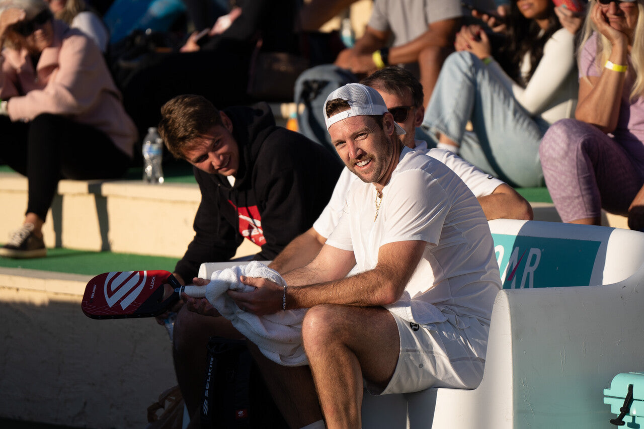 Pickleball pros Collin Shick and Jack Sock sit on a bench during a timeout in their match. Jack wipes his hands and pickleball grip on a sweat towel. 