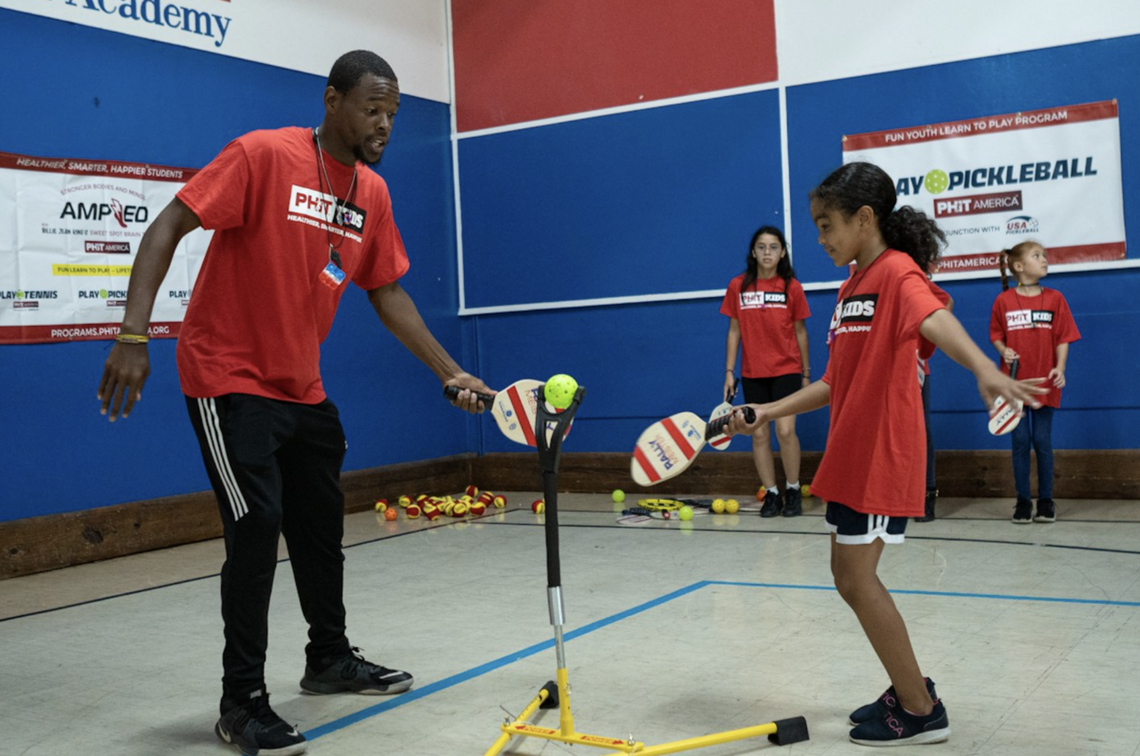 A young female student hits a pickleball off a tee while a male instructor looks on during a PHIT America pickleball course in a school gymnasium. 