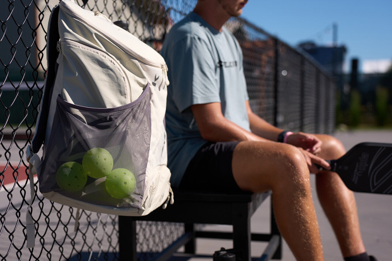 A white Selkirk backpack is clipped to a fence next to an outdoor pickleball court. A male pickleball player sits on a bench next to the bag. 