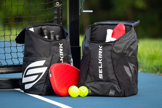 Two large Selkirk pickleball backpacks sit on a pickleball court. They are open to show they can store many paddles, balls, and other accessories. 