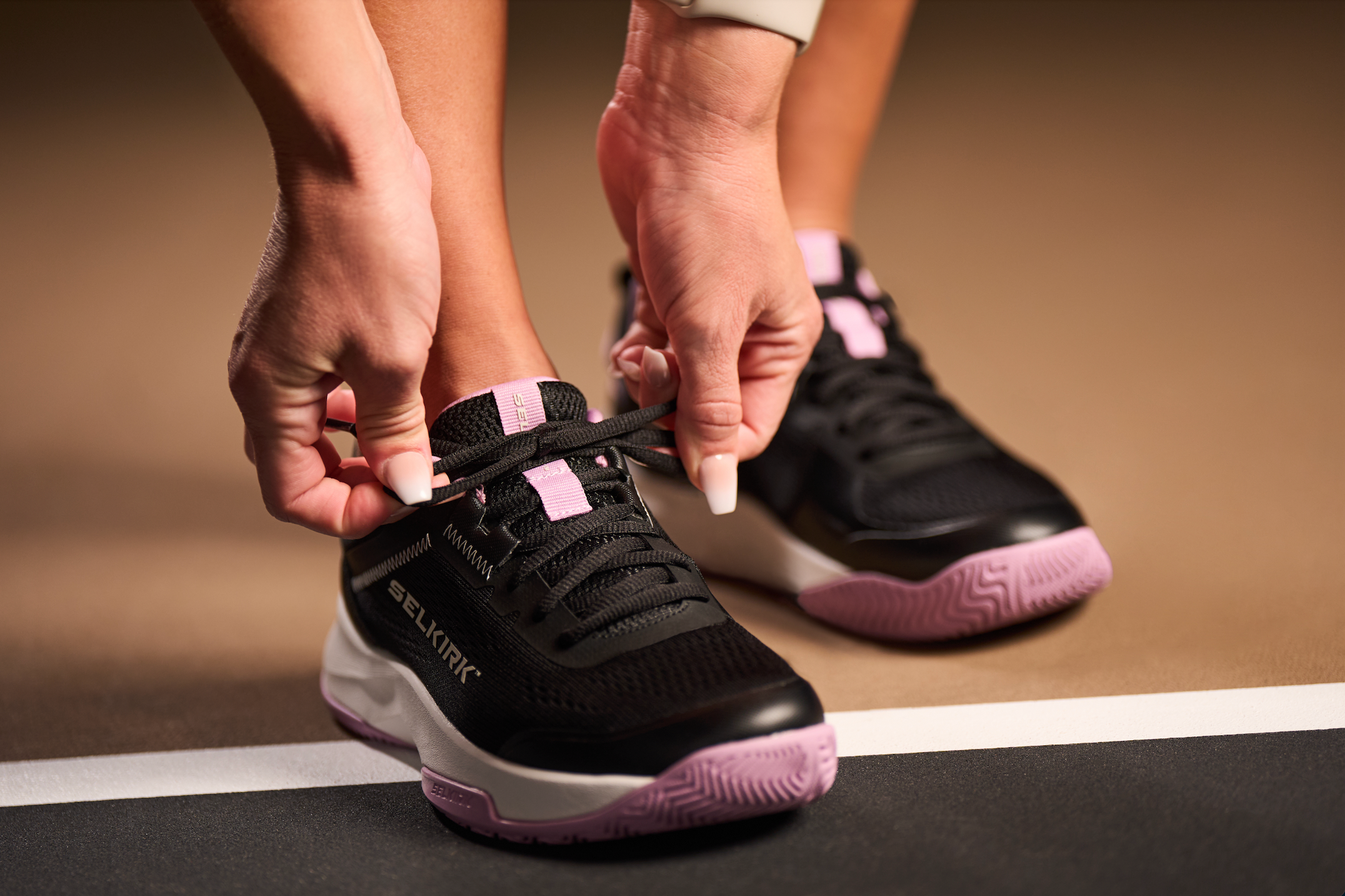 A close-up shot shoes a woman's hands as she ties her Selkirk pickleball court shoe on an indoor court.