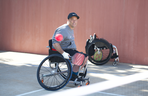 A man uses a wheelchair to play pickleball outdoors. He is smiling as he prepares to hit a ball. 