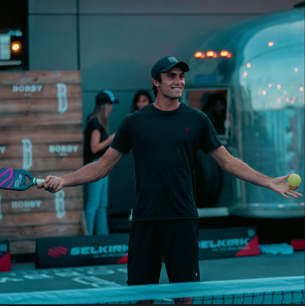 James Ignatowich pumps his arms in the air to get a crowd of spectators involved as he stands on an outdoor pickleball court. 