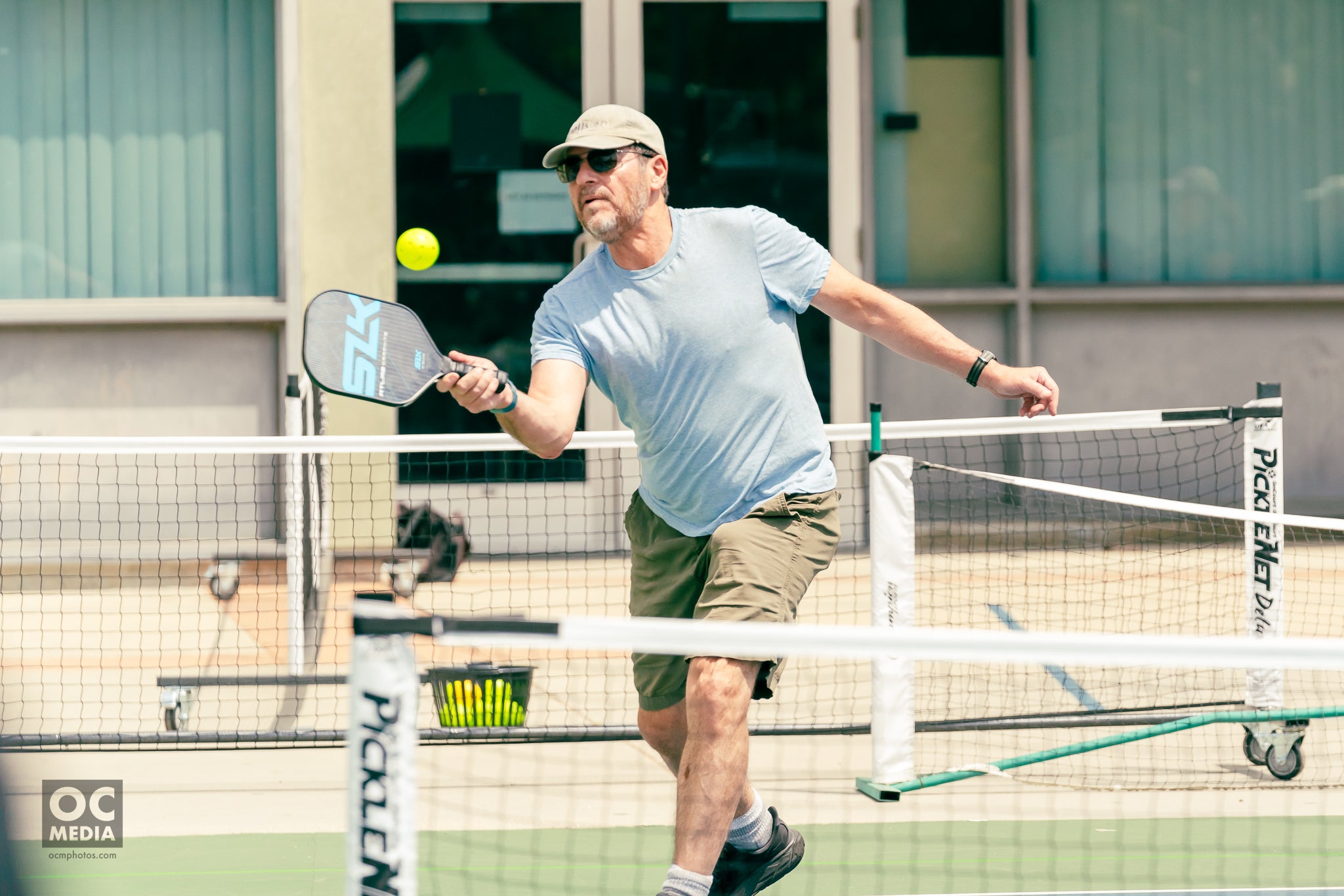 A man volleys a pickleball with a SLK paddle. He stands on the other side of a pickleball net, away from the camera. 