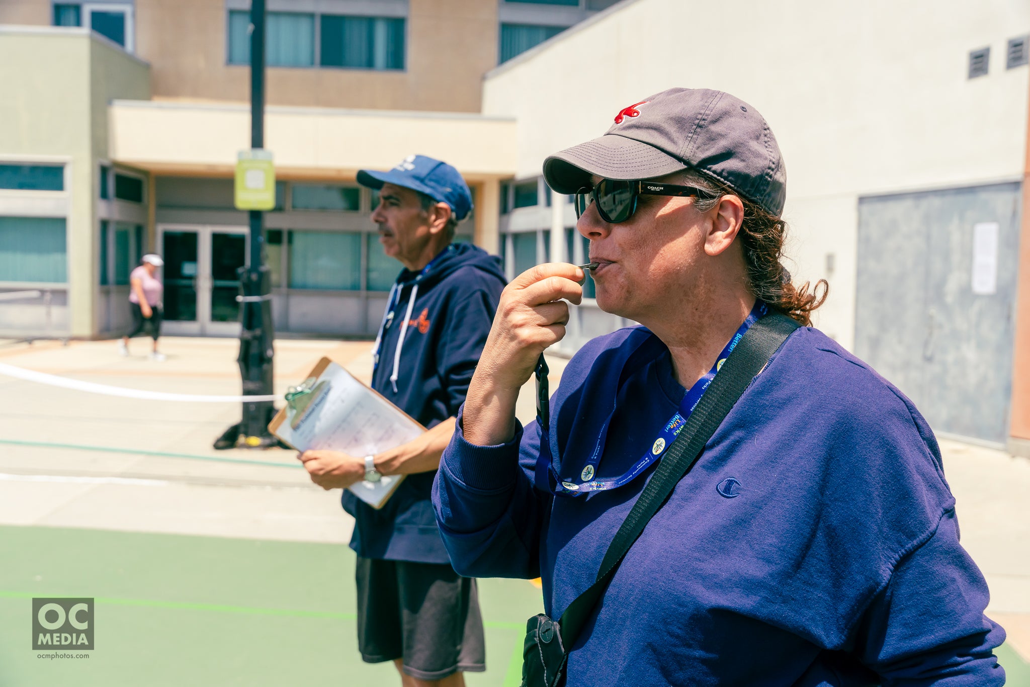 A woman close to the camera holds a whistle to her lips. In the background, a man holds a clipboard. 