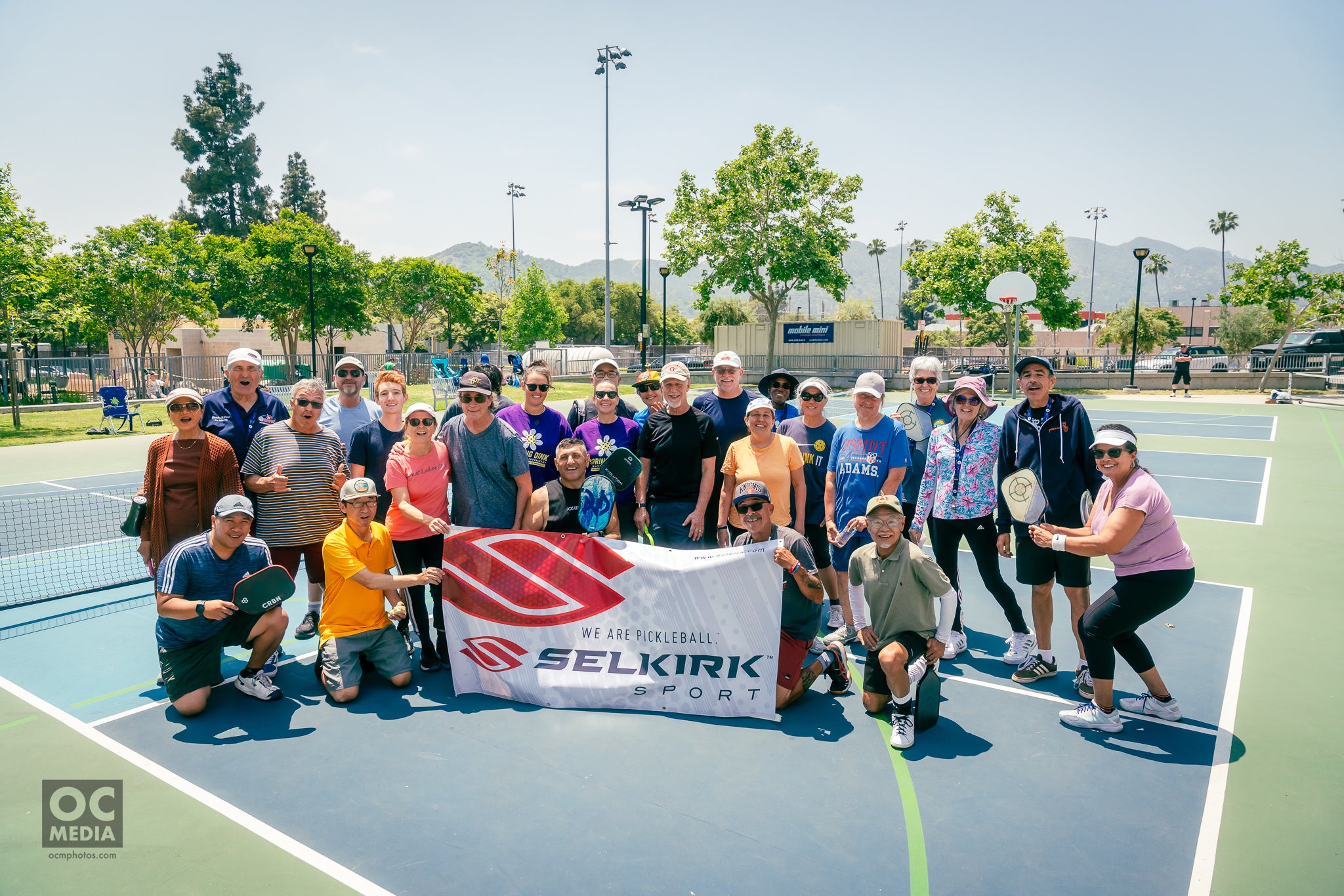 A group of pickleball players gather behind a Selkirk Sport banner. They are all standing on a pickleball court. In the background are mountains. 