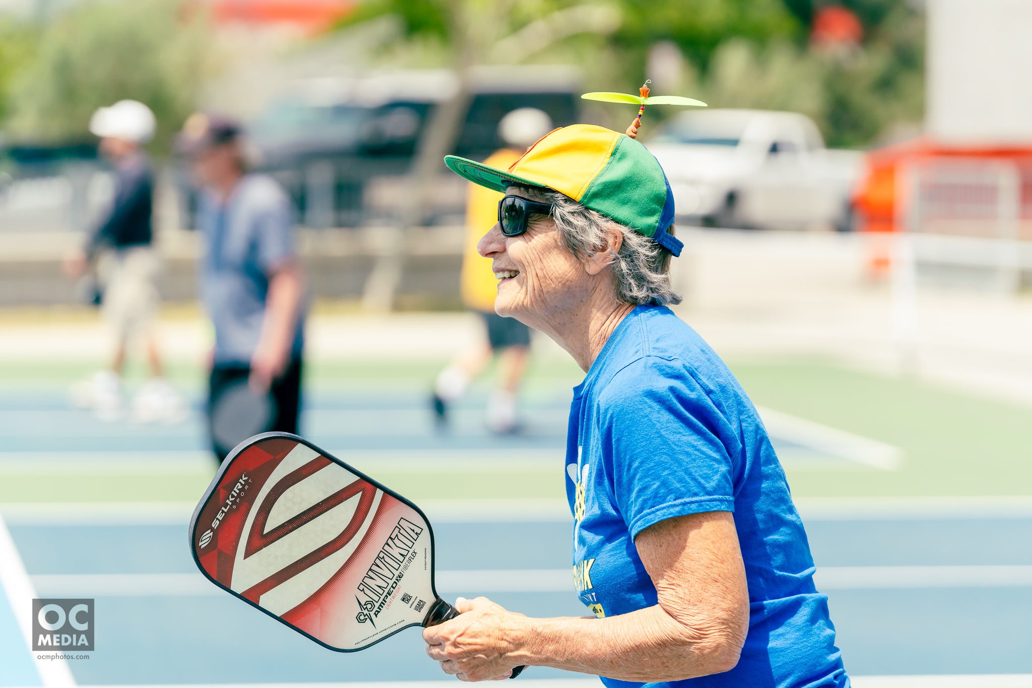 A woman holds a Selkirk paddle. She is smiling and wearing a baseball cap with a propellor on top. 