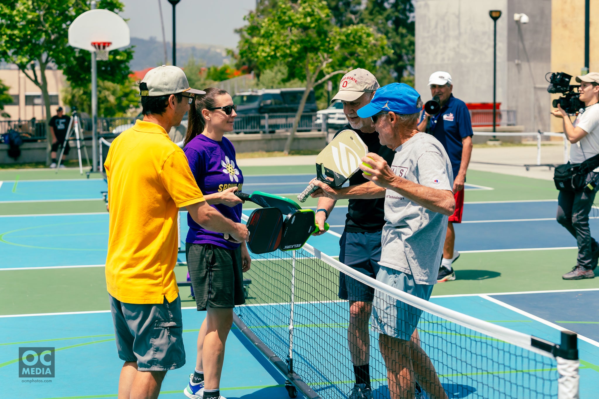 Four pickleball players tap Selkirk Paddles across a pickleball net. On one side is a young man and woman, on the the other side are two older men. 