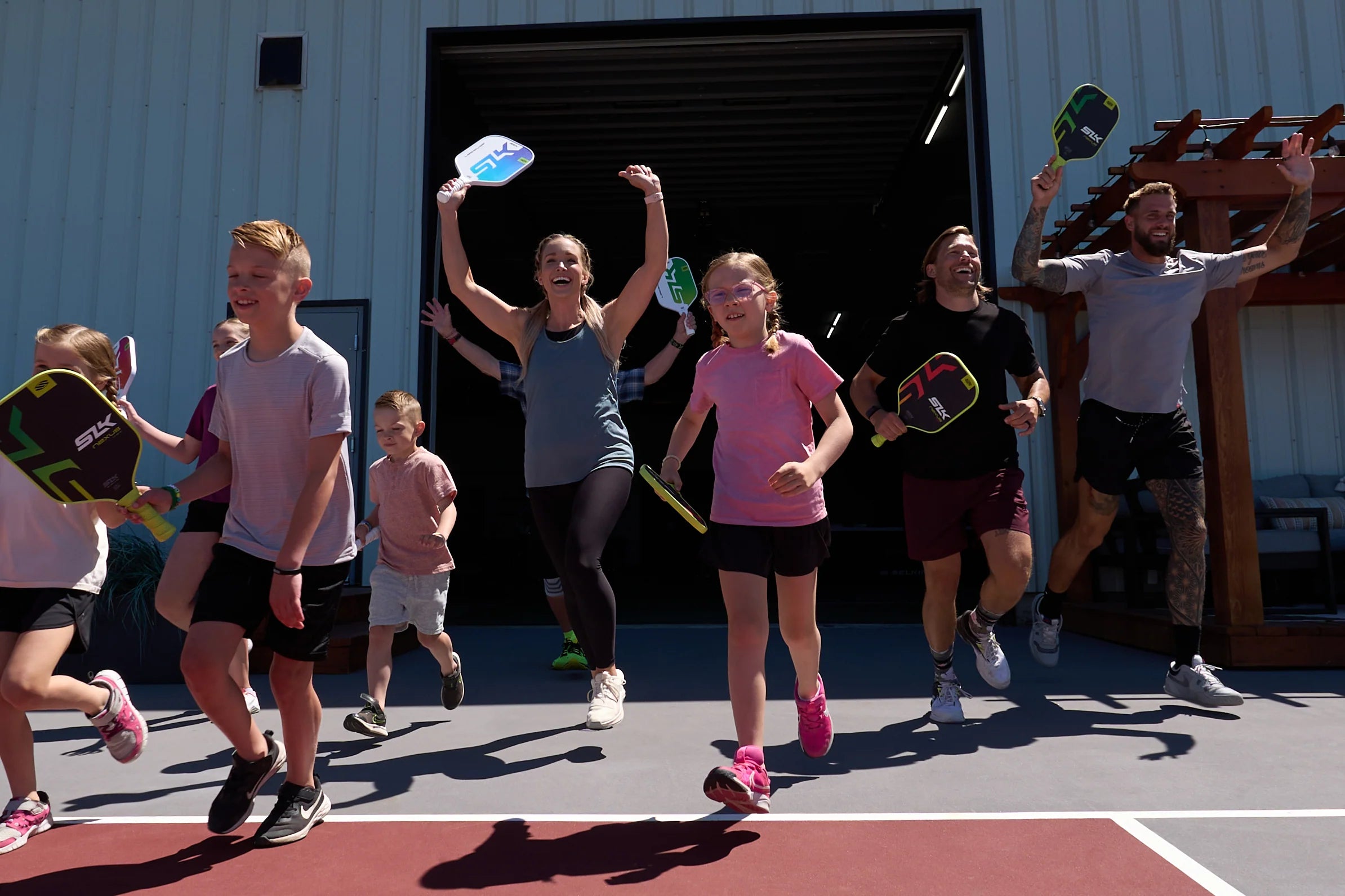 Group of children and adults joyfully playing pickleball with paddles outside.